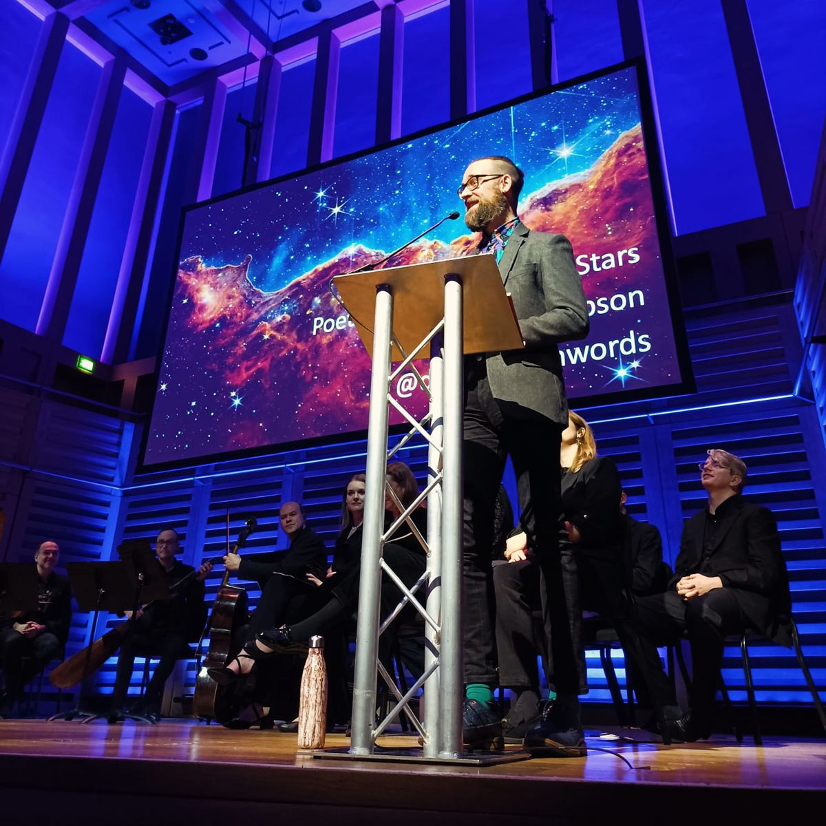 Dan reading at a lectern on stage with the Orchestra of the Age of Enlightenment, a projected star field behind him